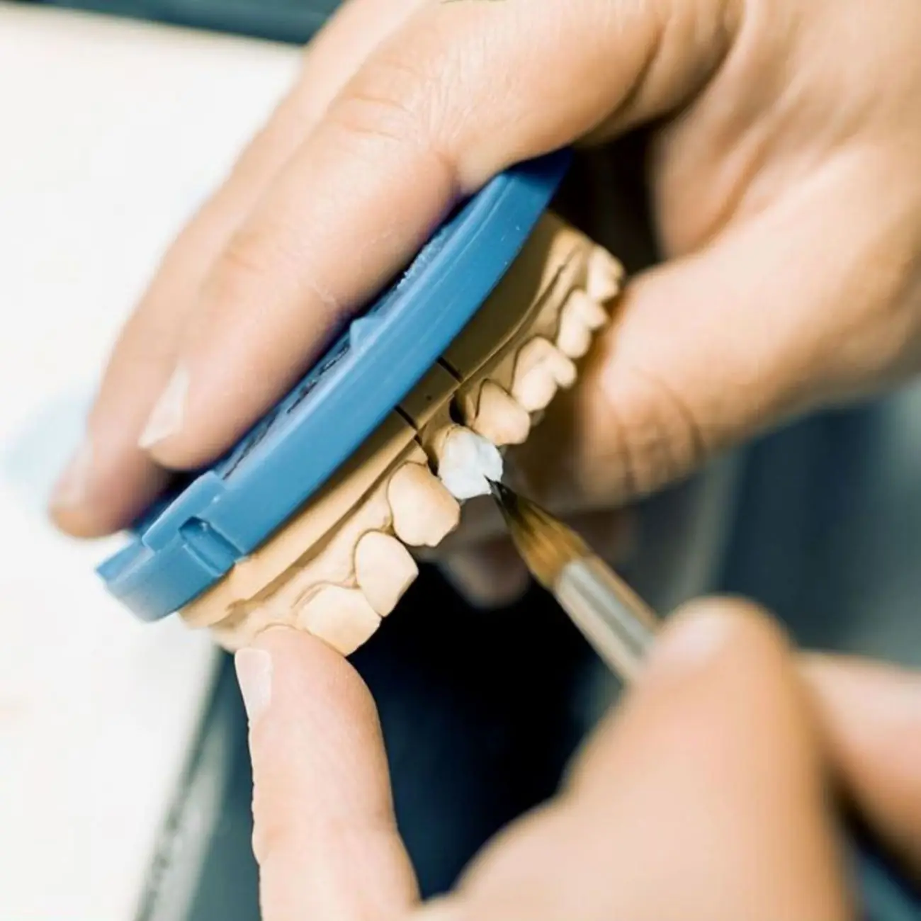 Mãos aplicando cerâmica com pincel fino sobre um modelo de gesso, dente por dente